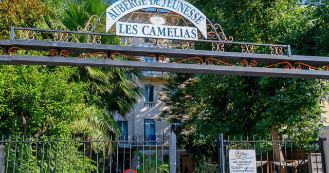 Während der Klassenfahrt nach Nizza übernachtet die Schulklasse in der Jugendherberge Les Camélias im Zentrum von Nizza. Auf dem Bild sehen wir den Eingan der Herberge hinter Bäumen, Palmen und einem Zaun.